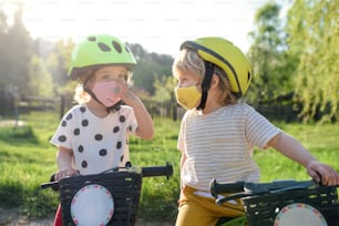 Bambino piccolo ragazzo e ragazza con maschere per il viso che giocano all'aperto con la bicicletta, concetto di coronavirus.