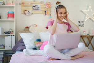 Cheerful young girl with headphones and laptop on bed, relaxing during quarantine.