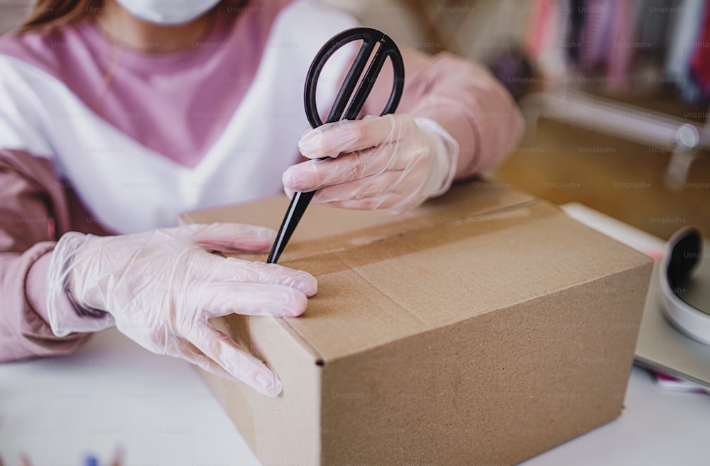 Young female student with face mask and gloves at the table, opening parcel box.