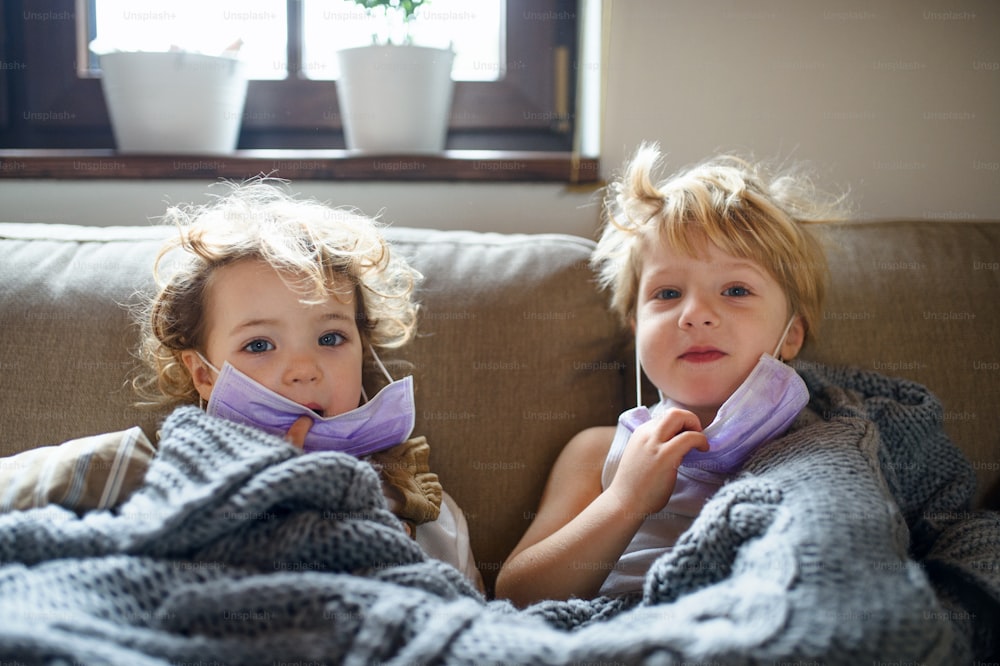 Dos niños pequeños enfermos con mascarilla en casa acostados en la cama, mirando a la cámara.