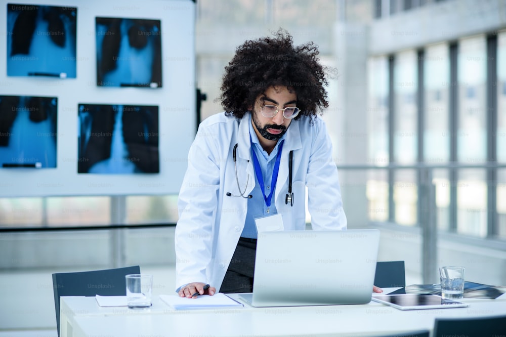 Retrato frontal de un médico serio parado en el hospital, usando una computadora portátil.
