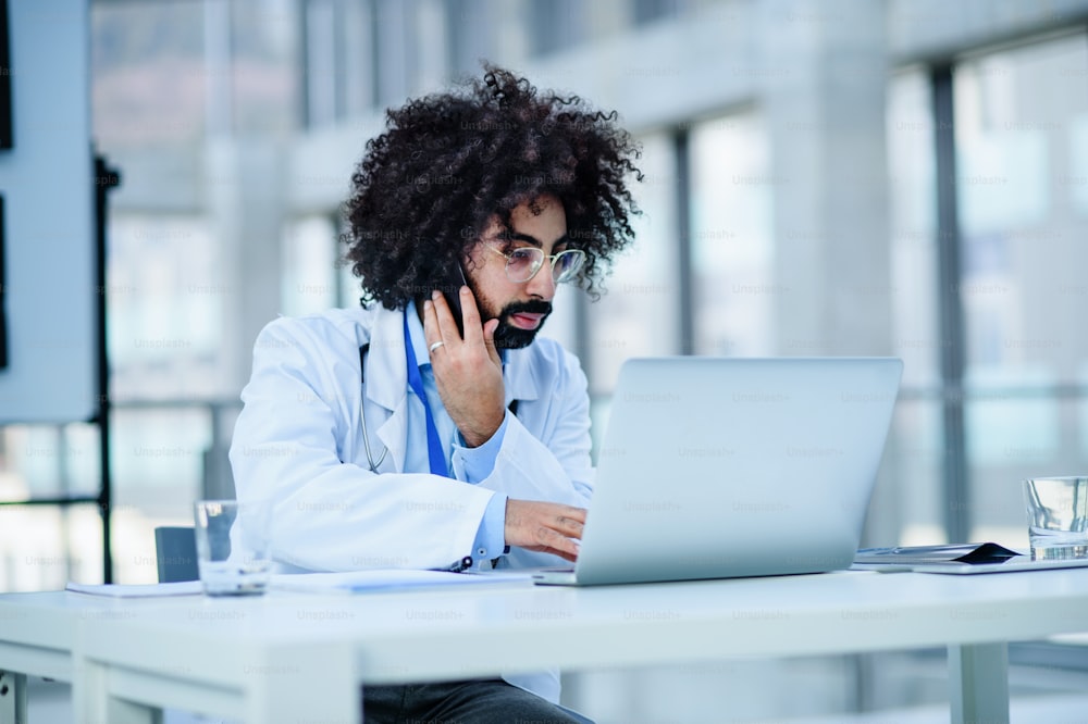 Porträt eines vielbeschäftigten männlichen Arztes, der im Krankenhaus sitzt und Laptop und Smartphone benutzt.