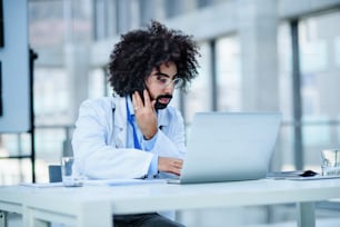 Portrait of busy male doctor sitting in hospital, using laptop and smartphone.