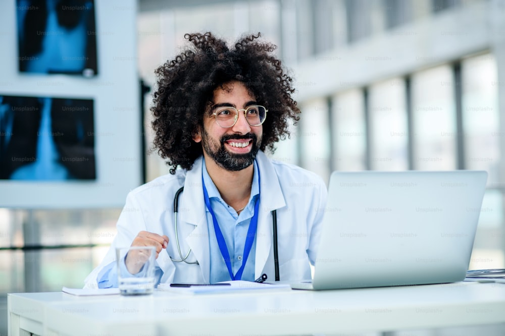 Retrato do médico masculino alegre e feliz sentado no hospital, usando laptop.