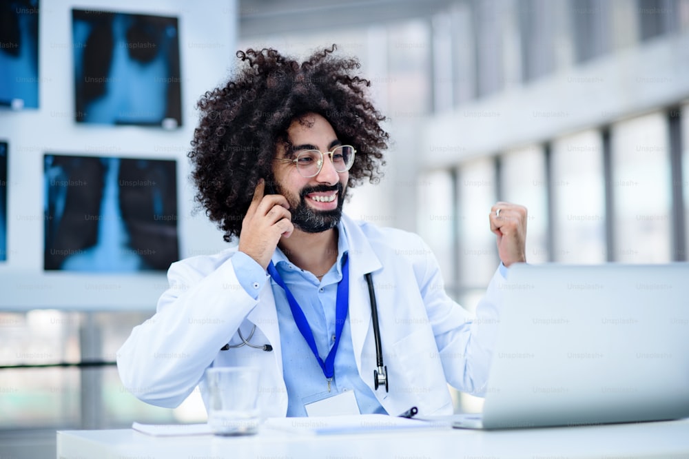Portrait d’un médecin masculin joyeux assis à l’hôpital, utilisant un ordinateur portable et un smartphone.