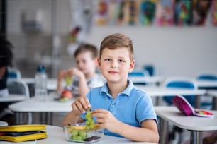 Ein kleiner glücklicher Schuljunge, der am Schreibtisch im Klassenzimmer sitzt und Traubenfrüchte isst.