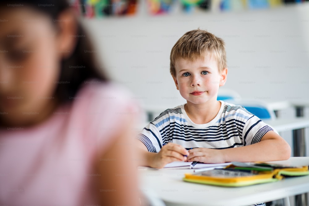 Um pequeno garoto feliz sentado na mesa da sala de aula, olhando para a câmera.