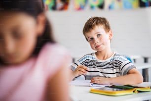 Um pequeno garoto feliz sentado na mesa da sala de aula, escrevendo e ouvindo o professor.