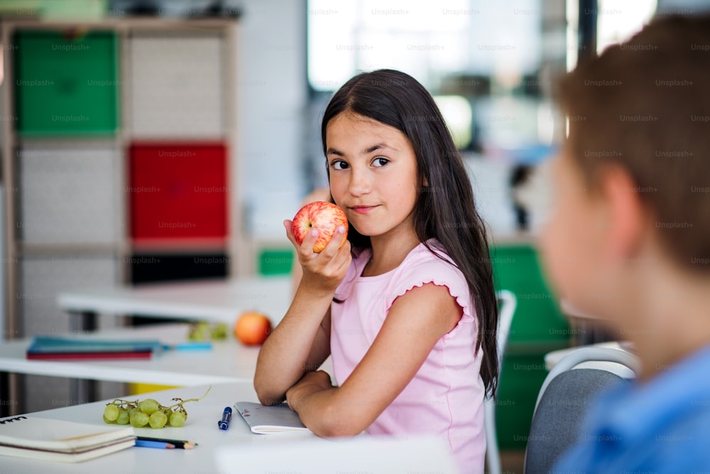 A small happy school girl sitting at the desk in classroom, eating apple fruit.