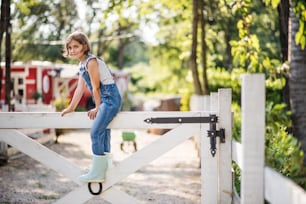 A portrait of happy small girl on family farm, sitting on wooden gate.