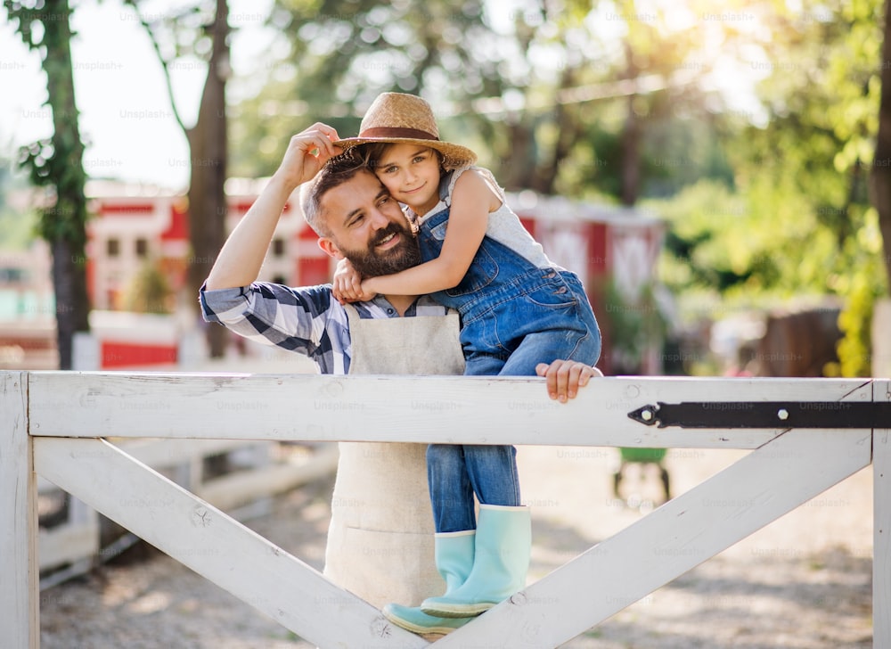 A portrait of father with small daughter outdoors on family farm, standing by wooden gate.