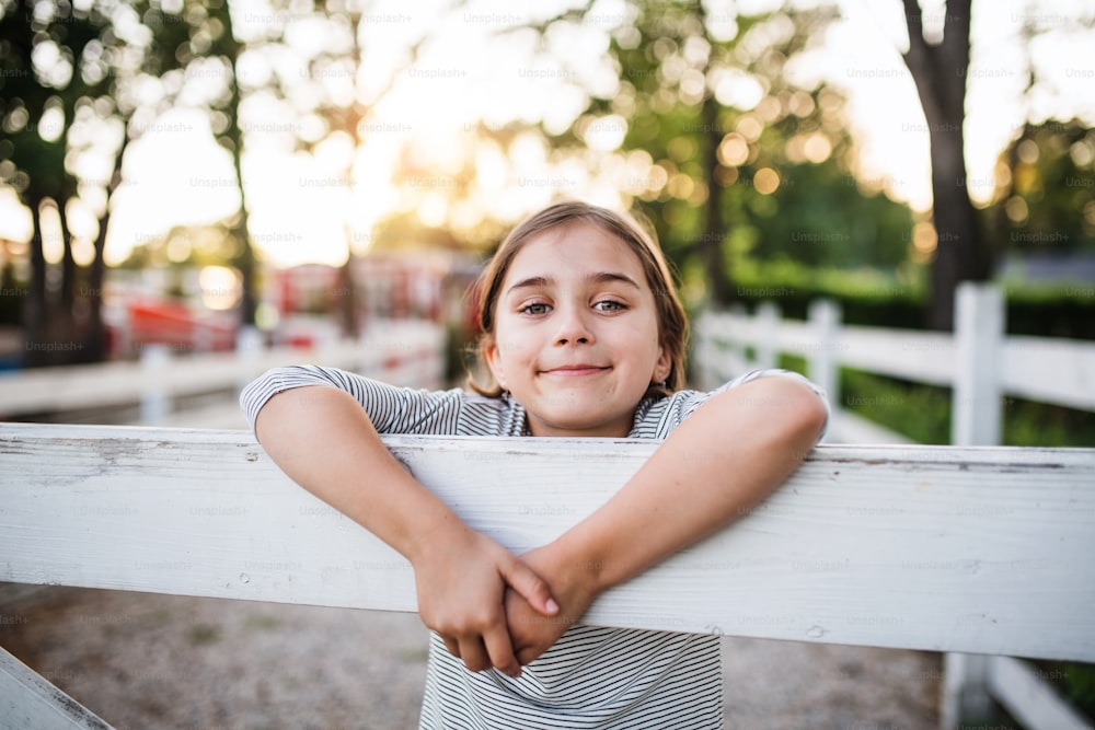 Ein Porträt eines glücklichen kleinen Mädchens auf dem Bauernhof der Familie, das am Holztor steht.