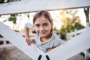 Ein Porträt eines glücklichen kleinen Mädchens auf dem Bauernhof der Familie, das am Holztor steht.