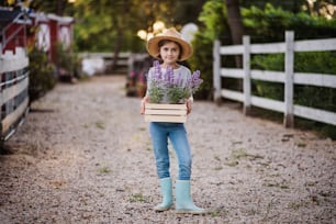 Eine Vorderansicht eines kleinen Mädchens mit Hut, das draußen auf dem Familienbauernhof steht und Pflanzen hält.