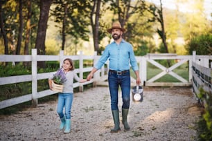 성숙한 아버지와 고무 장화를 신은 작은 딸이 손을 잡고 작은 가족 농장에서 야외를 걷고 있다.