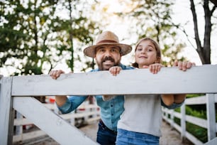 Un retrato de un padre con una hija pequeña al aire libre en la granja familiar, mirando a la cámara.
