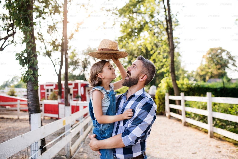Un padre feliz con una hija pequeña parada al aire libre en la granja familiar, jugando con sombrero.