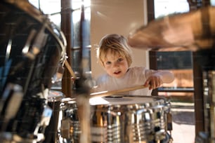 Front view of happy small boy indoors at home, playing drums.