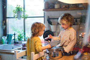 Heureux petit garçon et fille à l’intérieur dans la cuisine à la maison, aidant à cuisiner.
