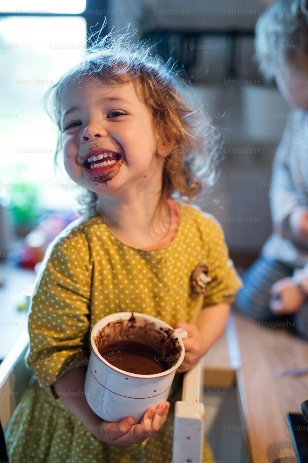 Glückliches kleines Mädchen mit schmutzigem Mund drinnen in der Küche zu Hause, Pudding essend.