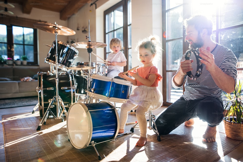 Portrait of happy small children with father indoors at home, playing drums.