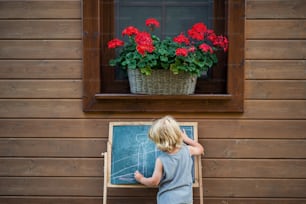 A rear view of happy small blond boy playing outdoors by house in summer.