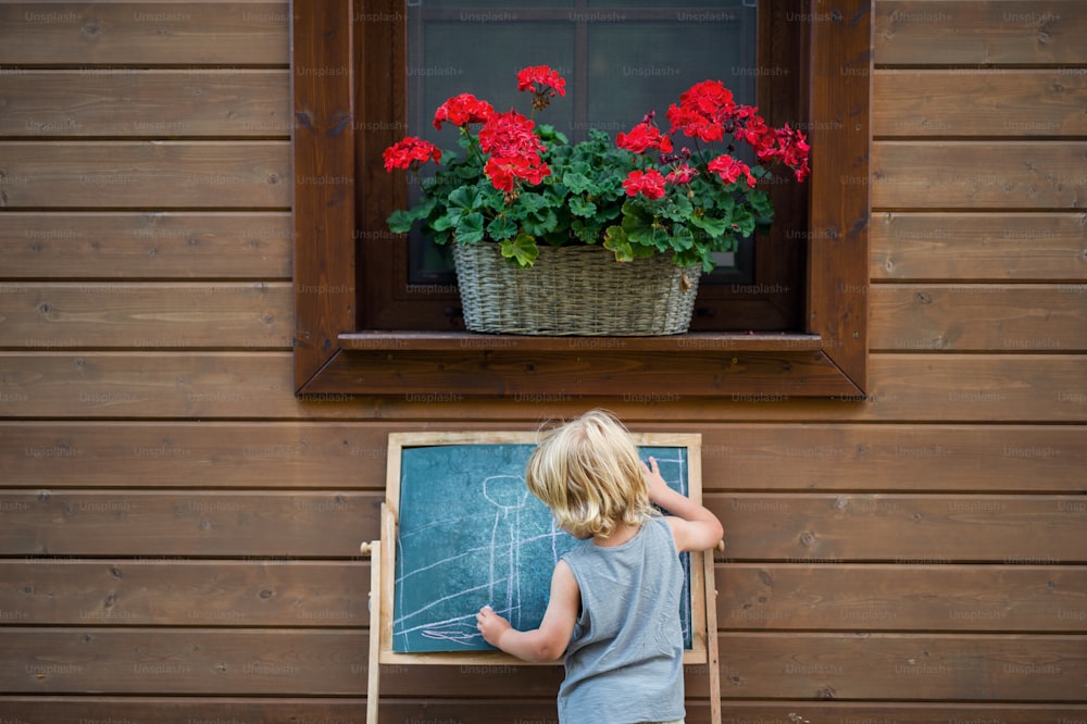 Eine Rückansicht eines glücklichen kleinen blonden Jungen, der im Sommer im Freien am Haus spielt.