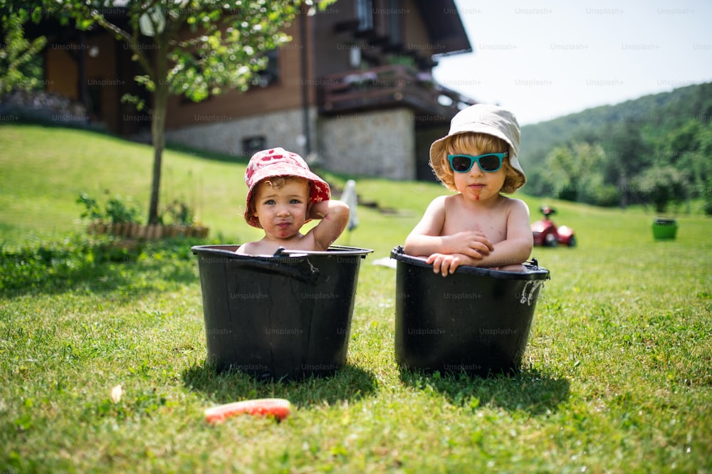 Topless small boy and girl with hats in buckets outdoors in summer garden, looking at camera.