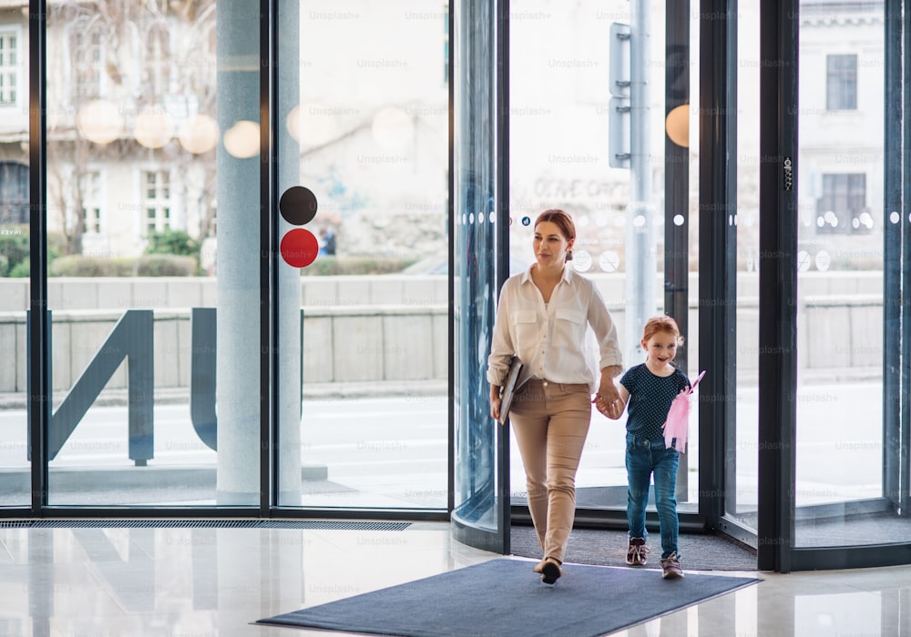 Eine Geschäftsfrau mit kleiner Tochter betritt ein Bürogebäude, geht und hält Händchen.