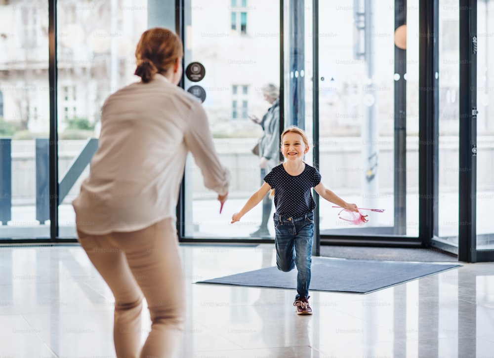 Una vista trasera de una mujer de negocios saludando a una pequeña hija feliz en un edificio de oficinas.