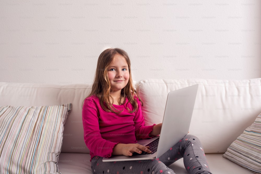 Little girl sitting on sofa with a laptop computer at home. Happy child playing indoors using PC.