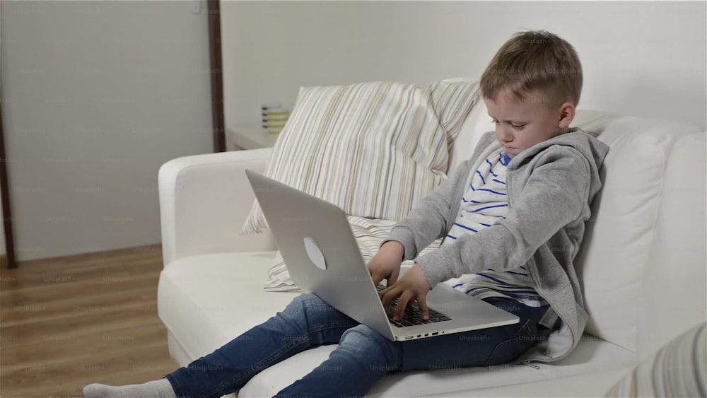 어린 소년은 집에서 노트북 컴퓨터를 들고 소파에 앉아 있다. PC를 사용하여 실내에서 노는 행복한 아이.