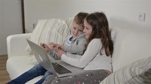 Niña y niño sentados en el sofá con una computadora portátil en casa. Niños felices jugando en interiores usando PC.