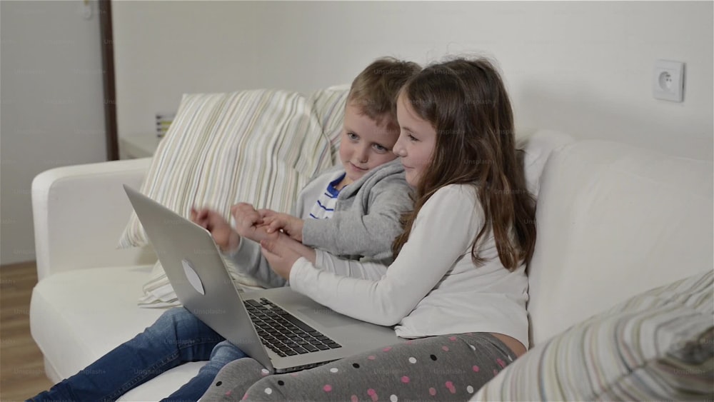 Menina e menino sentados no sofá com um computador portátil em casa. Crianças felizes brincando dentro de casa usando o PC.