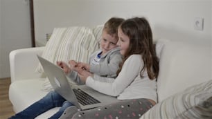 Little girl and boy sitting on sofa with a laptop computer at home. Happy children playing indoors using PC.