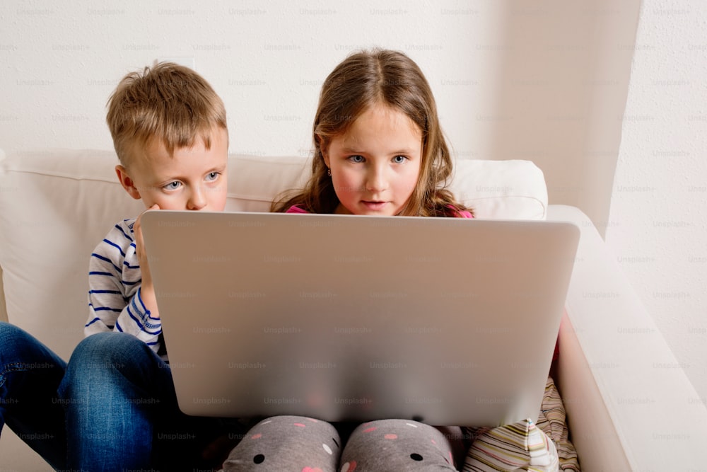 Petite fille et petit garçon assis sur un canapé avec un ordinateur portable à la maison. Des enfants heureux jouent à l’intérieur en utilisant un PC.