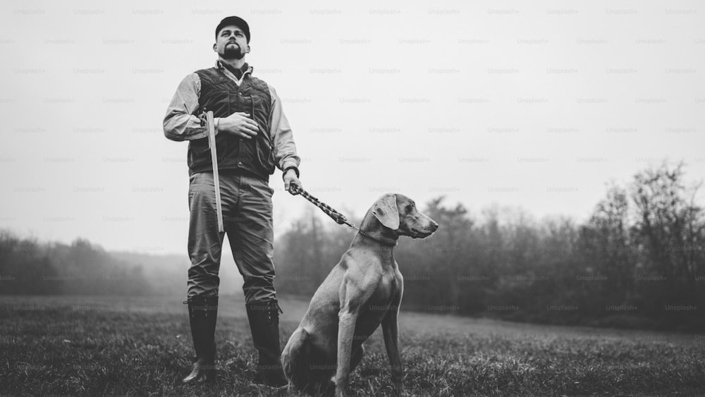 Un cacciatore con un cane in abiti da tiro tradizionali sul campo con fucile da caccia, foto in bianco e nero.