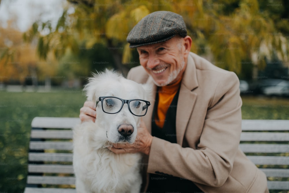 A happy senior man looking at camera and embracing his dog wearing glasses on bench outdoors in city.