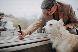 Un uomo anziano felice seduto sulla panchina e scattando selfie con il suo cane all'aperto in città.