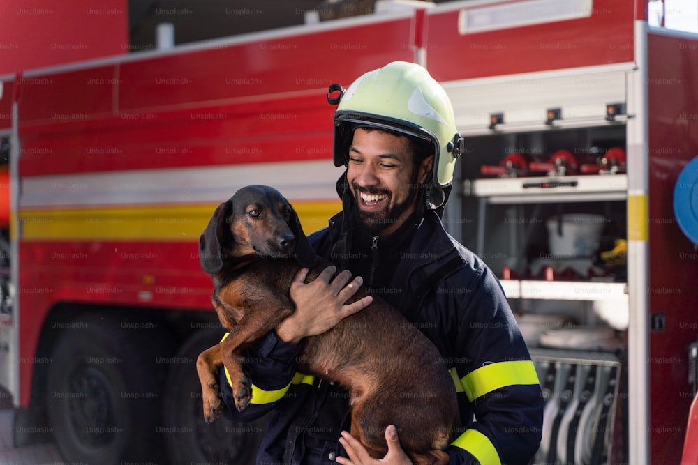 Ein glücklicher reifer Feuerwehrmann, der einen Hund hält und mit einem Feuerwehrauto im Hintergrund in die Kamera schaut