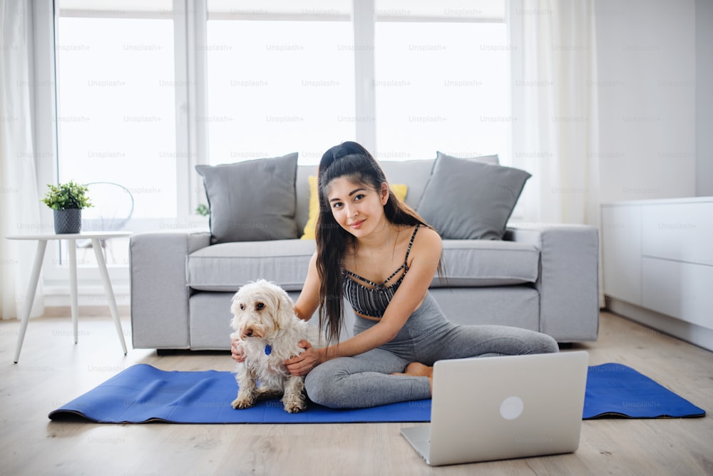 Eine junge Sportlerin mit Laptop und Hund, die zu Hause drinnen Sport treibt und in die Kamera schaut.