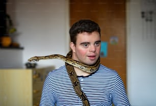 Portrait of down syndrome adult man sitting indoors in bedroom at home, playing with pet snake.