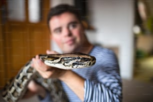 Ritratto di uomo adulto con sindrome di Down seduto in casa in camera da letto a casa, giocando con il serpente domestico.