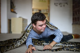 Ritratto di uomo adulto con sindrome di Down seduto in casa in camera da letto a casa, giocando con il serpente domestico.