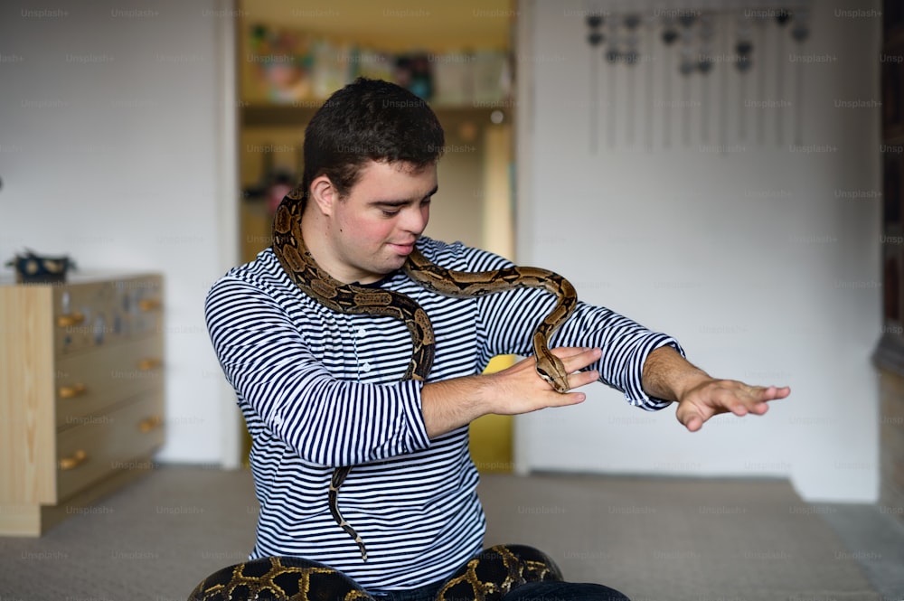Retrato de un hombre adulto con síndrome de Down sentado en el interior del dormitorio de su casa, jugando con una serpiente mascota.