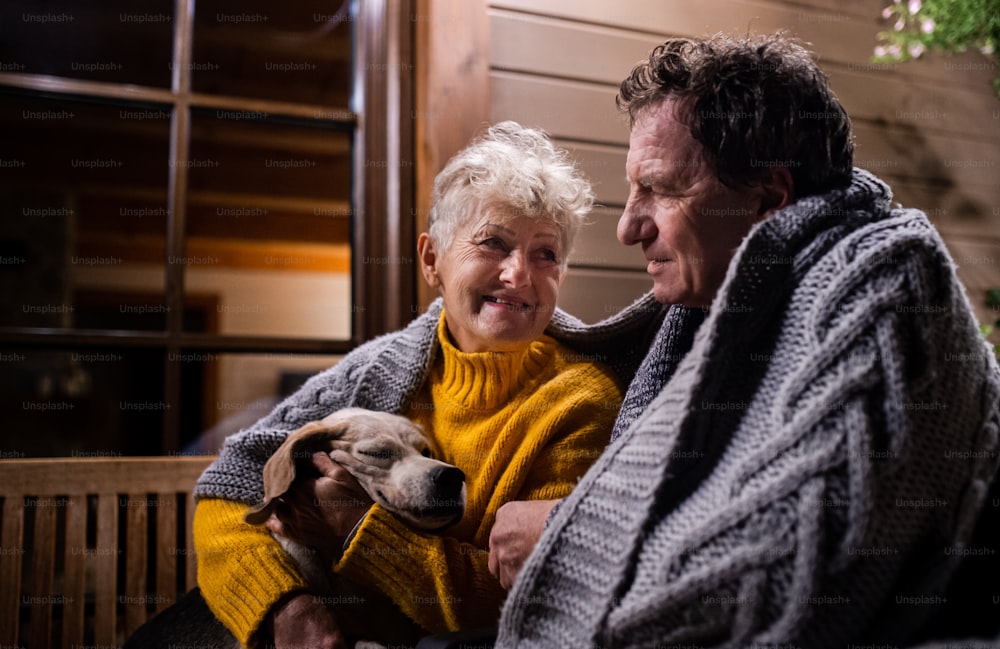 Retrato de una pareja mayor con un perro sentado por la noche en la terraza envuelto en una manta, hablando.