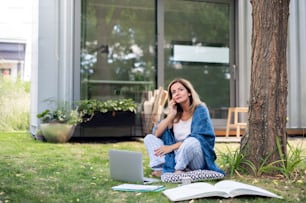 Reife Frau im Pyjama, die im Homeoffice im Freien auf Gras im Garten arbeitet, mit Smartphone.