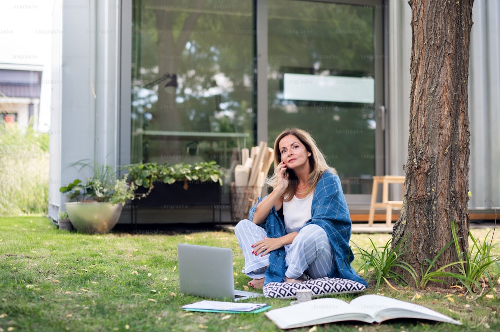 Mulher madura de pijama trabalhando em home office ao ar livre na grama no jardim, usando smartphone.
