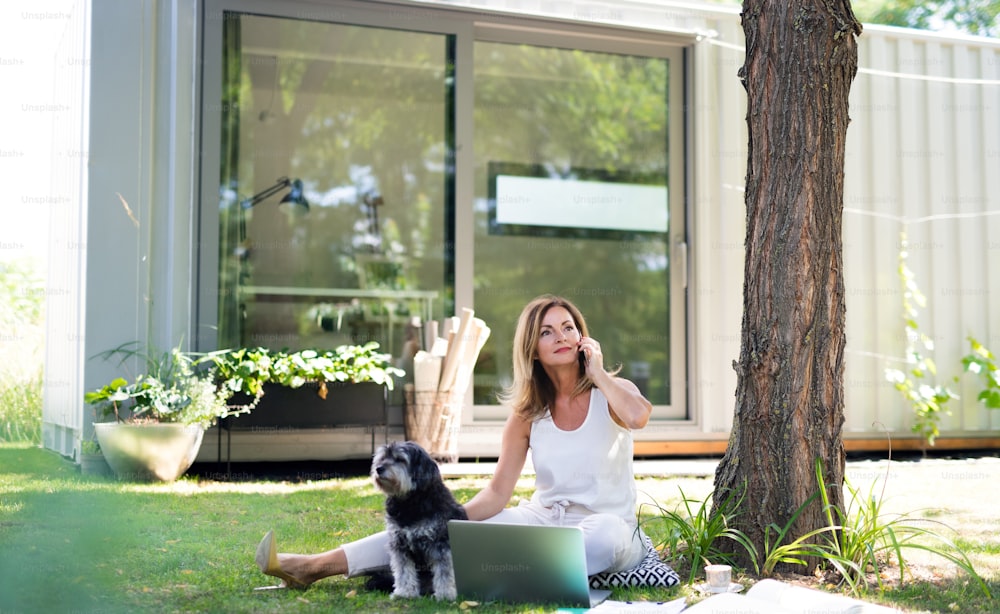 Mulher madura com cão de estimação trabalhando em home office ao ar livre no jardim, usando laptop.