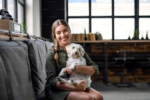 Portrait d’une jeune femme d’affaires avec un chien assis à l’intérieur dans le bureau, regardant la caméra.